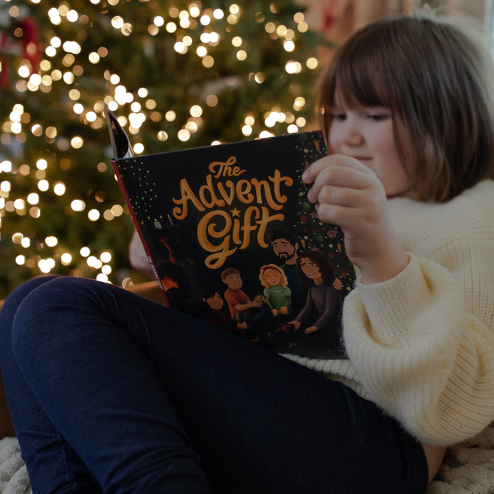 Child reading 'The Advent Gift' book in front of a decorated Christmas tree.