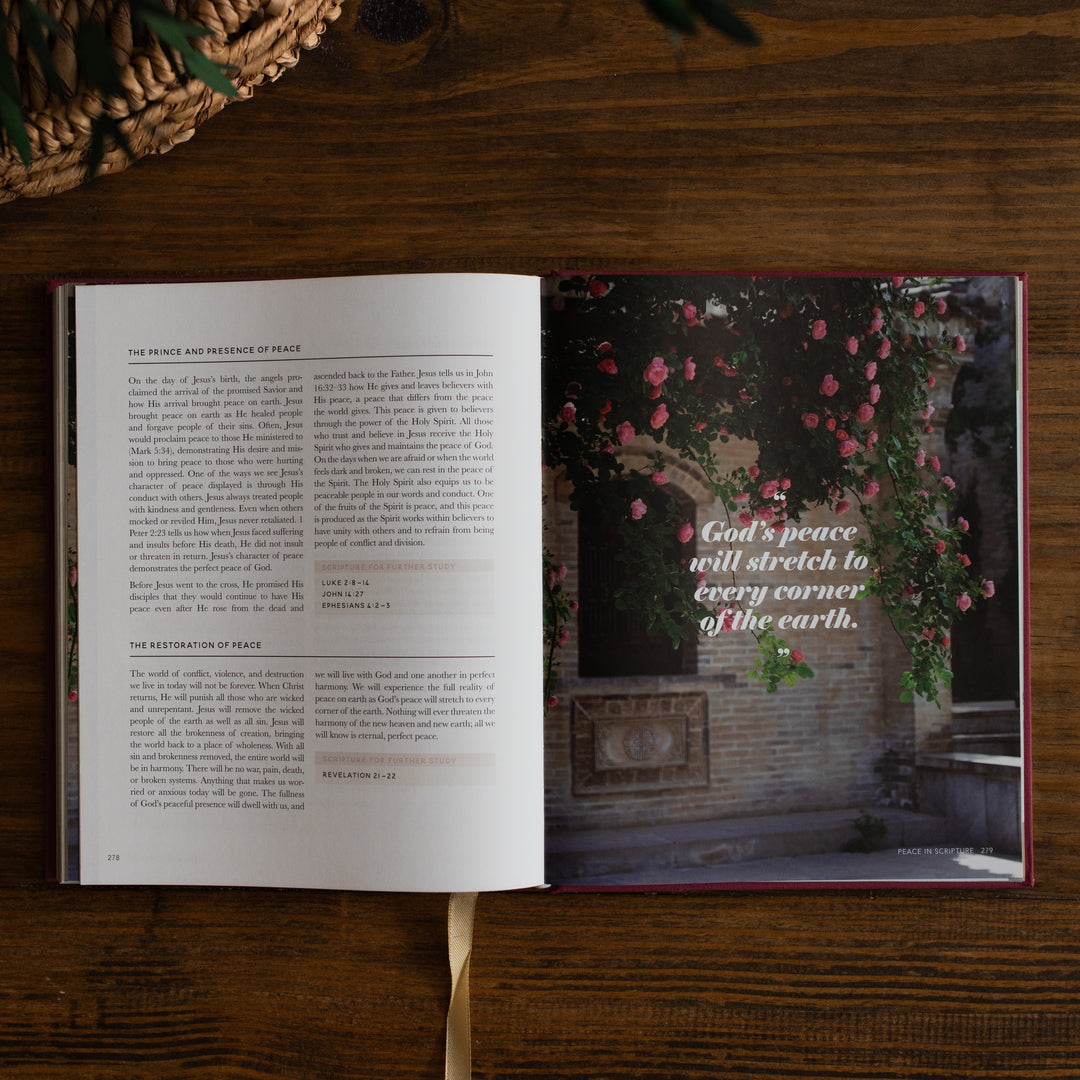 An open book laid flat on a dark wooden table, showing printed text on the left page and a scenic photograph with pink flowers and a quote on the right page. A woven basket and greenery are partially visible around it.