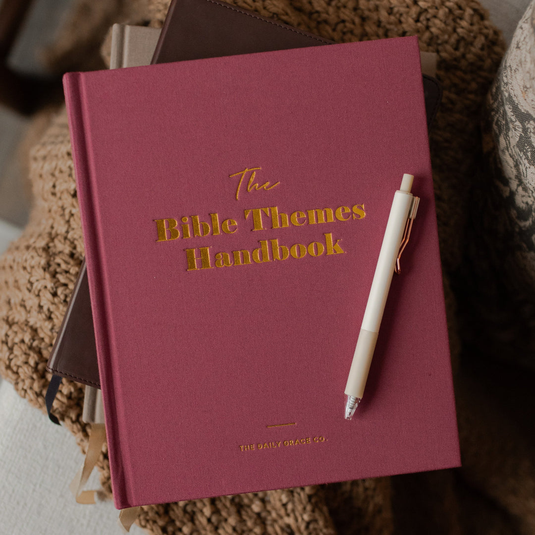 A dusty rose pink hardcover book titled 'The Bible Themes Handbook' with gold foil lettering, resting on a white armchair draped with a chunky brown knit blanket, with a white pen on top.