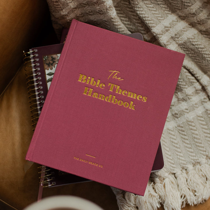 A burgundy hardcover book titled 'The Bible Themes Handbook' in gold lettering, styled on a brown leather couch with a beige plaid throw blanket and a cup of coffee in the foreground.