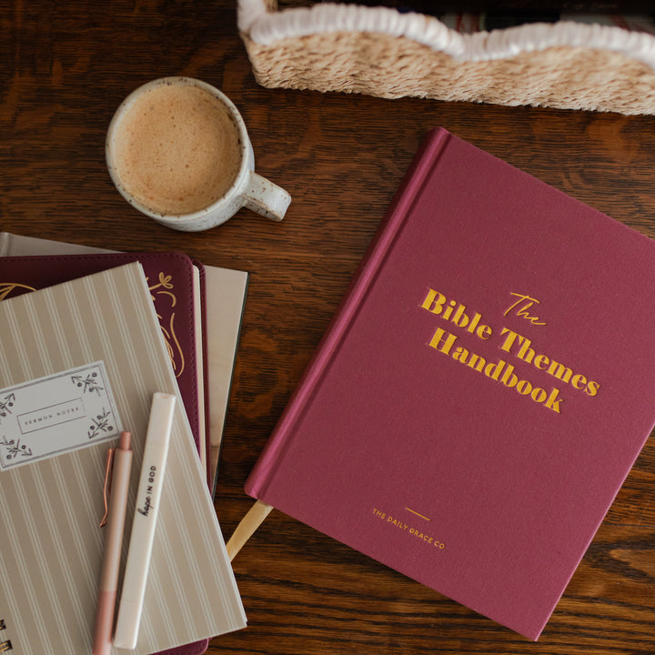 A styled flat-lay of 'The Bible Themes Handbook' by The Daily Prayer Co. on a dark wooden table, surrounded by a coffee cup, striped notebooks, and a pen.