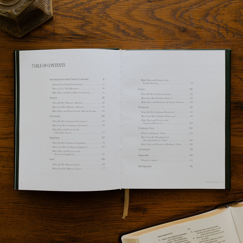 The Church Calendar opened to the table of contents page laying on wooden table next an open bible and a bronze candlestick