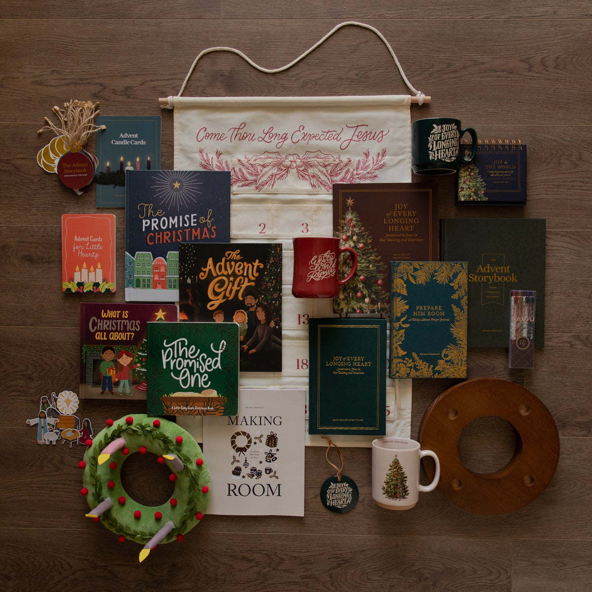 A styled flat-lay collection of Christmas-themed items arranged on a dark wood background, including a large cream fabric banner with red script, several books, mugs, a green wreath, a wooden ring, and small decorative pieces.