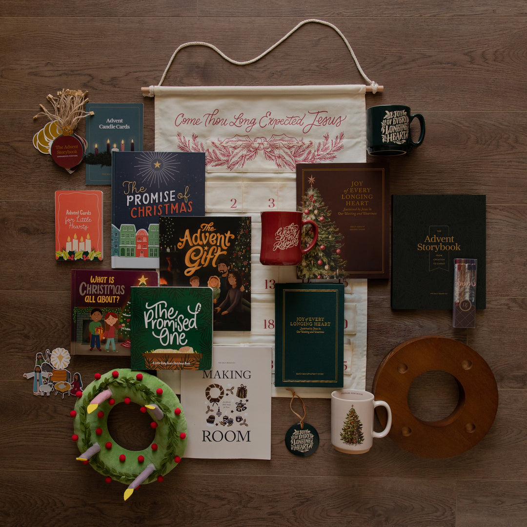 A styled flat-lay of various books, mugs, a wall hanging canvas, a green plush wreath, and a wooden candle ring on a dark wood background.