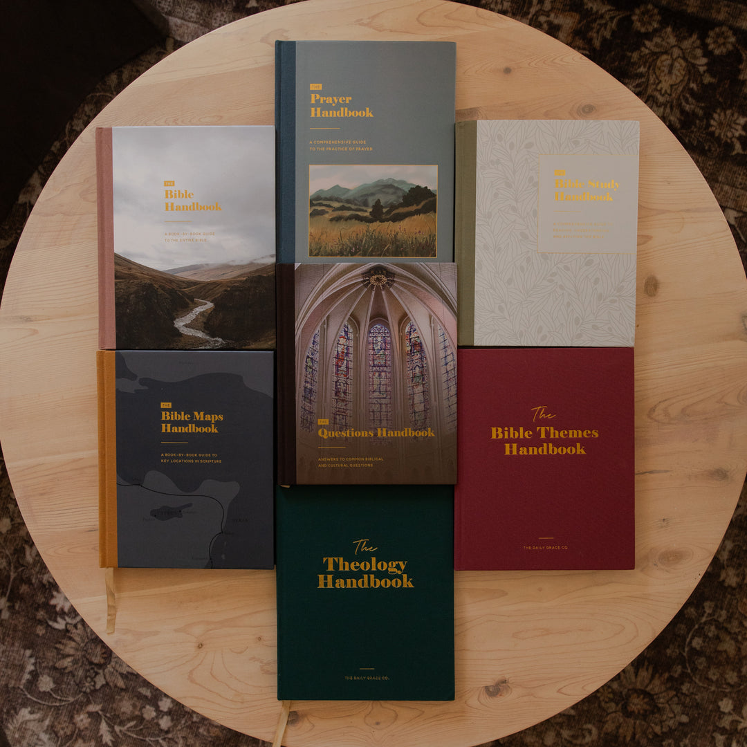 Seven handbooks from The Daily Grace Co. are arranged in a grid on a round wooden table. The books feature various colored covers including gray, burgundy, green, and beige, with gold lettering.