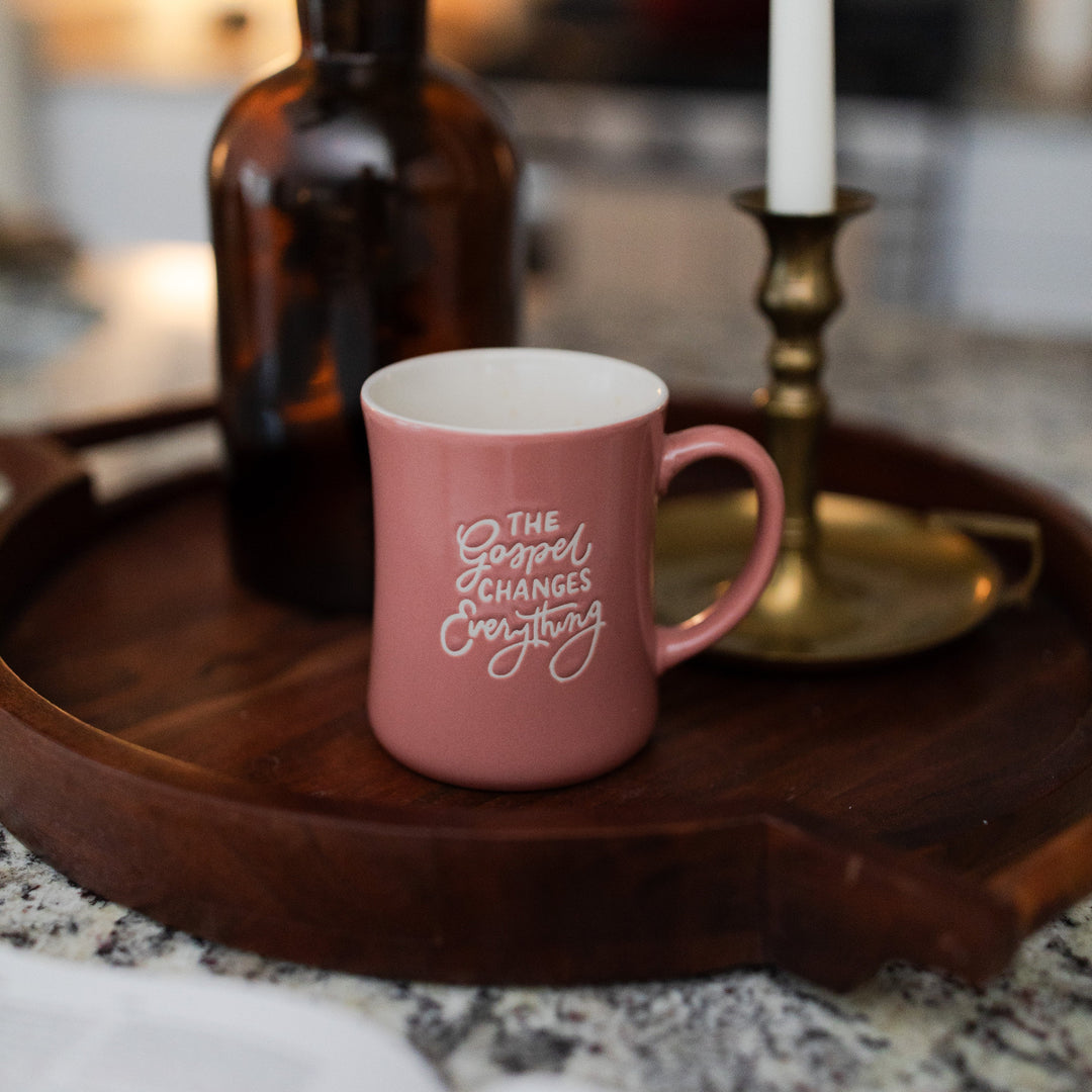 The Gospel Changes Everything Primrose Mug siting on a wooden round tray with a bronze candle and a brown glass bottle behind it