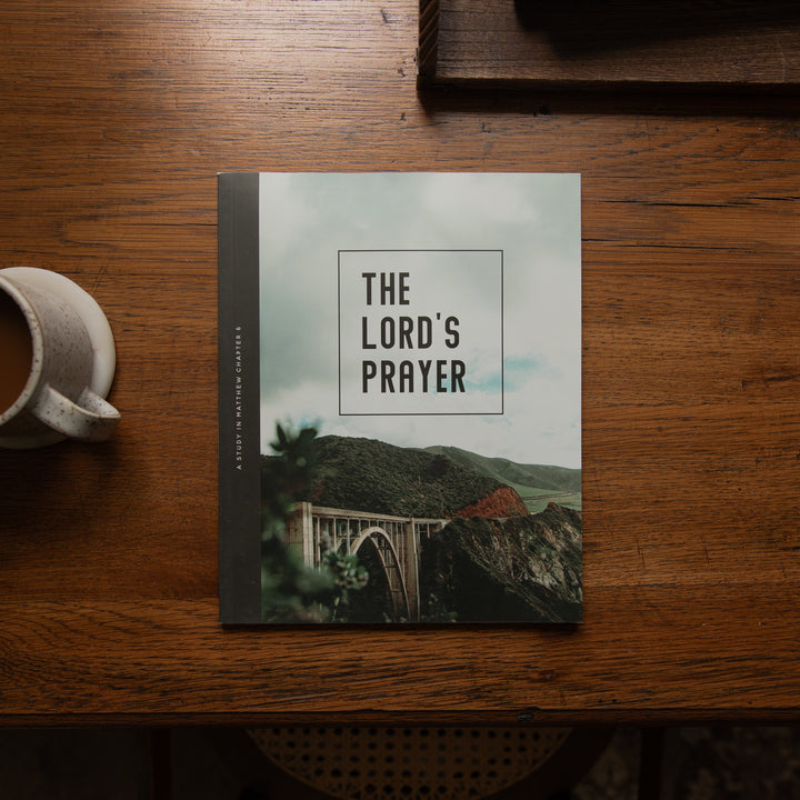 A book titled 'The Lord's Prayer' by Joshua Marshall lies open on a rustic wooden table, styled with a ceramic coffee mug on the left and a dark wooden tray in the background.