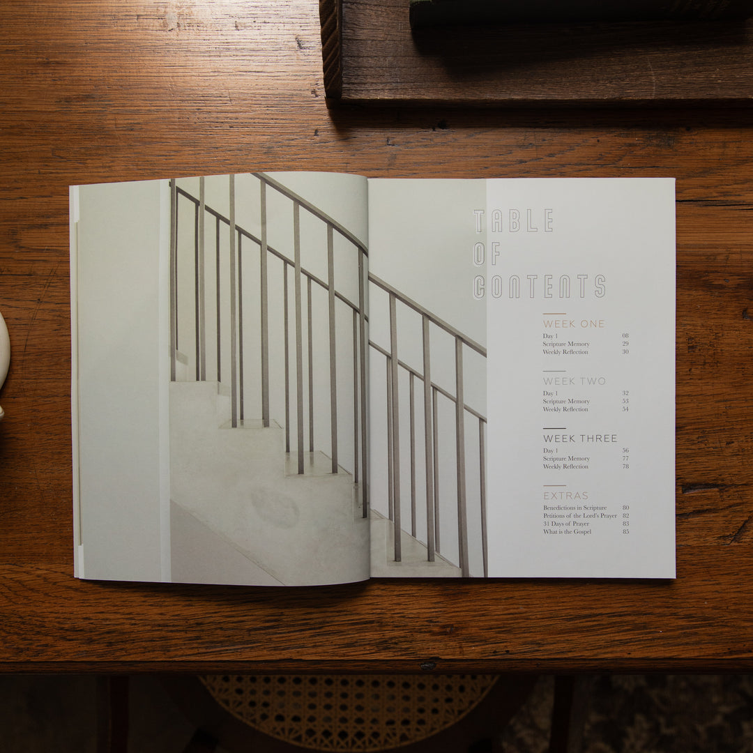 An open printed booklet or magazine lies on a dark wooden table, showing a staircase photograph on the left page and a table of contents on the right page.