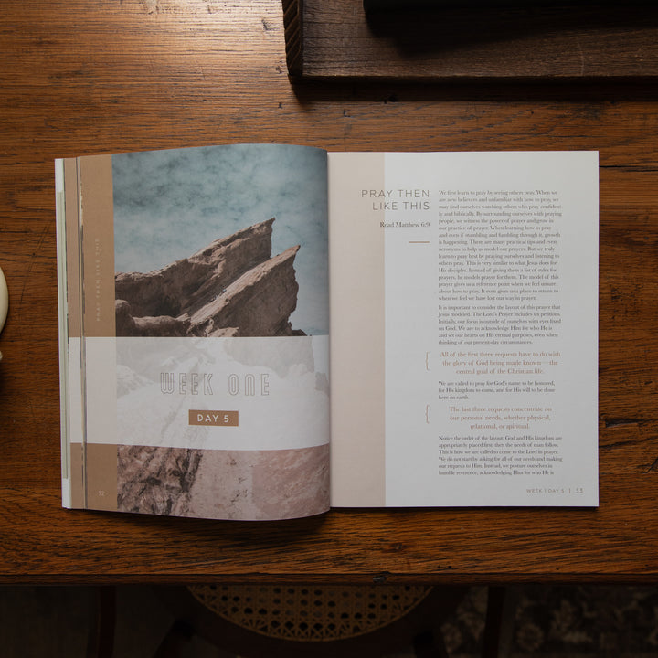 An open book or magazine laid flat on a wooden table, showing two printed pages with a desert rock landscape photo on the left page and a text passage on the right page. The cover has a visible spine band reading 'WEEK ONE DAY 1'.