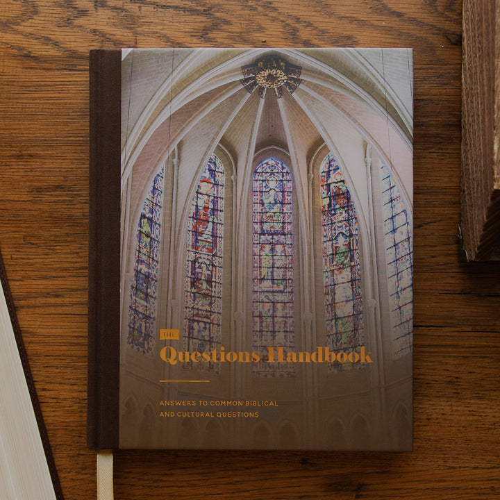 A hardcover book titled "Questions Handbook" lies on a wooden tabletop, with an open Bible at left and part of another book at right. The cover shows a photograph of a cathedral interior with stained glass windows and the title in gold text.