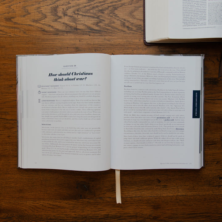 An open book laid flat on a wooden table, showing printed pages with the heading 'How should Christians think about war?' and a small dark blue bookmark tab on the right page. Another book is partially visible at the top right.