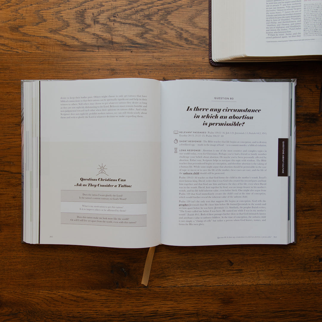 An open book laid flat on a wooden table, showing printed pages with text and a small bookmark hanging from the bottom center. Another book is partially visible at the top right.