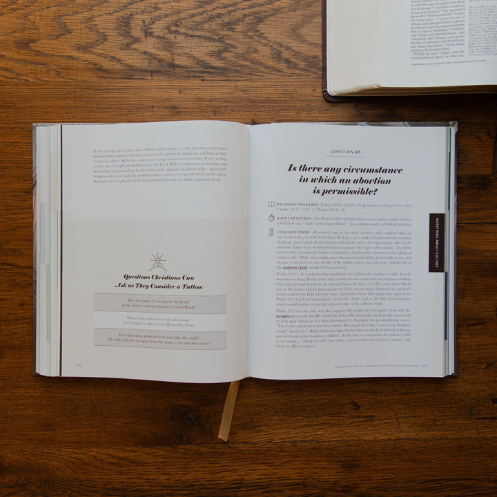 An open book laid flat on a wooden table, showing printed pages with text and a small bookmark hanging from the bottom center. Another book is partially visible at the top right.