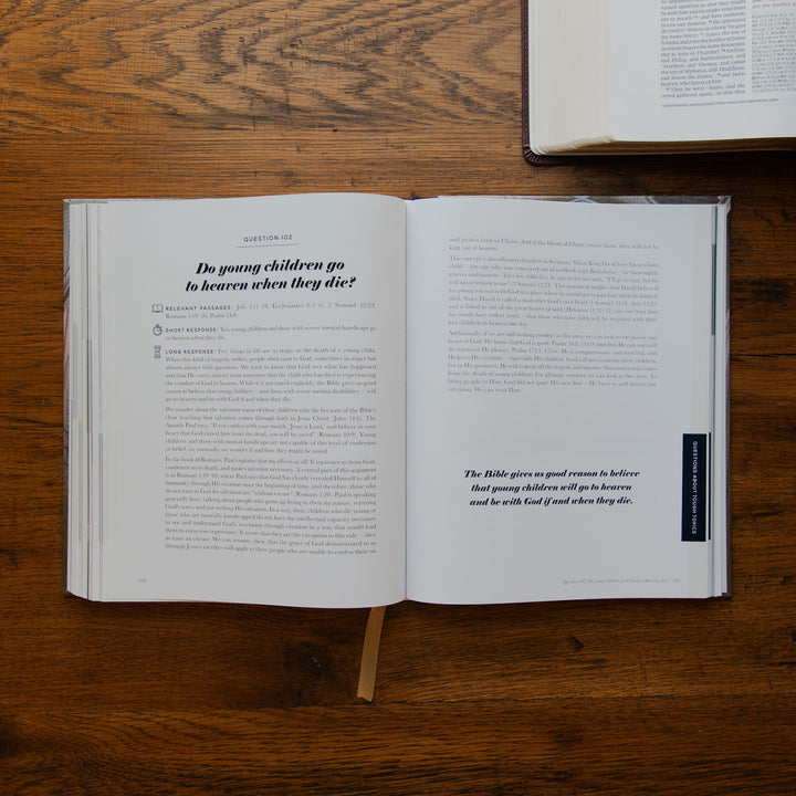 An open book laid flat on a wooden tabletop, showing printed text pages with a small dark vertical label on the right page. Another book is partially visible at the top right.