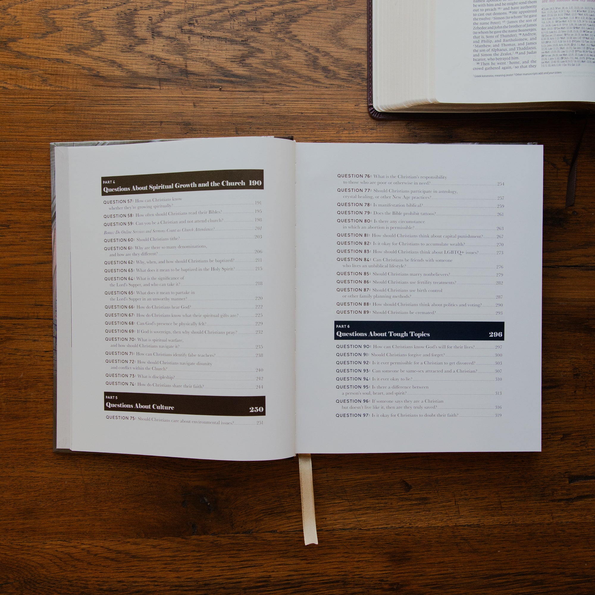 An open book showing printed pages with a table of contents, placed on a wooden tabletop. Another book is partially visible at the top right.