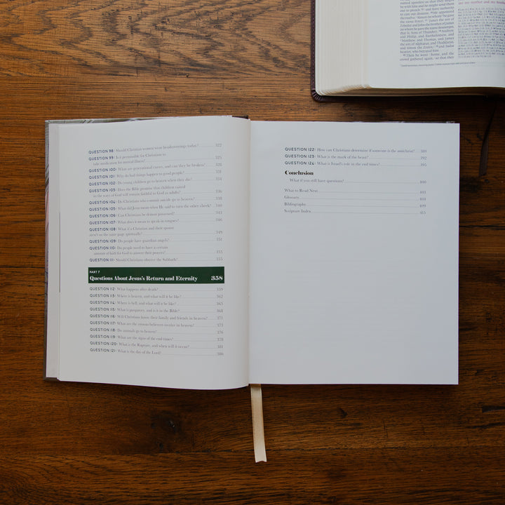 An open book showing printed text and a table of contents on a wooden tabletop, with another book partially visible at the top right.