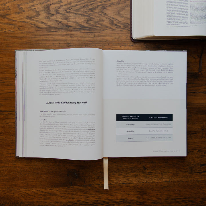 An open book laid flat on a wooden table, showing printed text pages with a bookmark ribbon extending downward. Another closed book is partially visible at the top right.