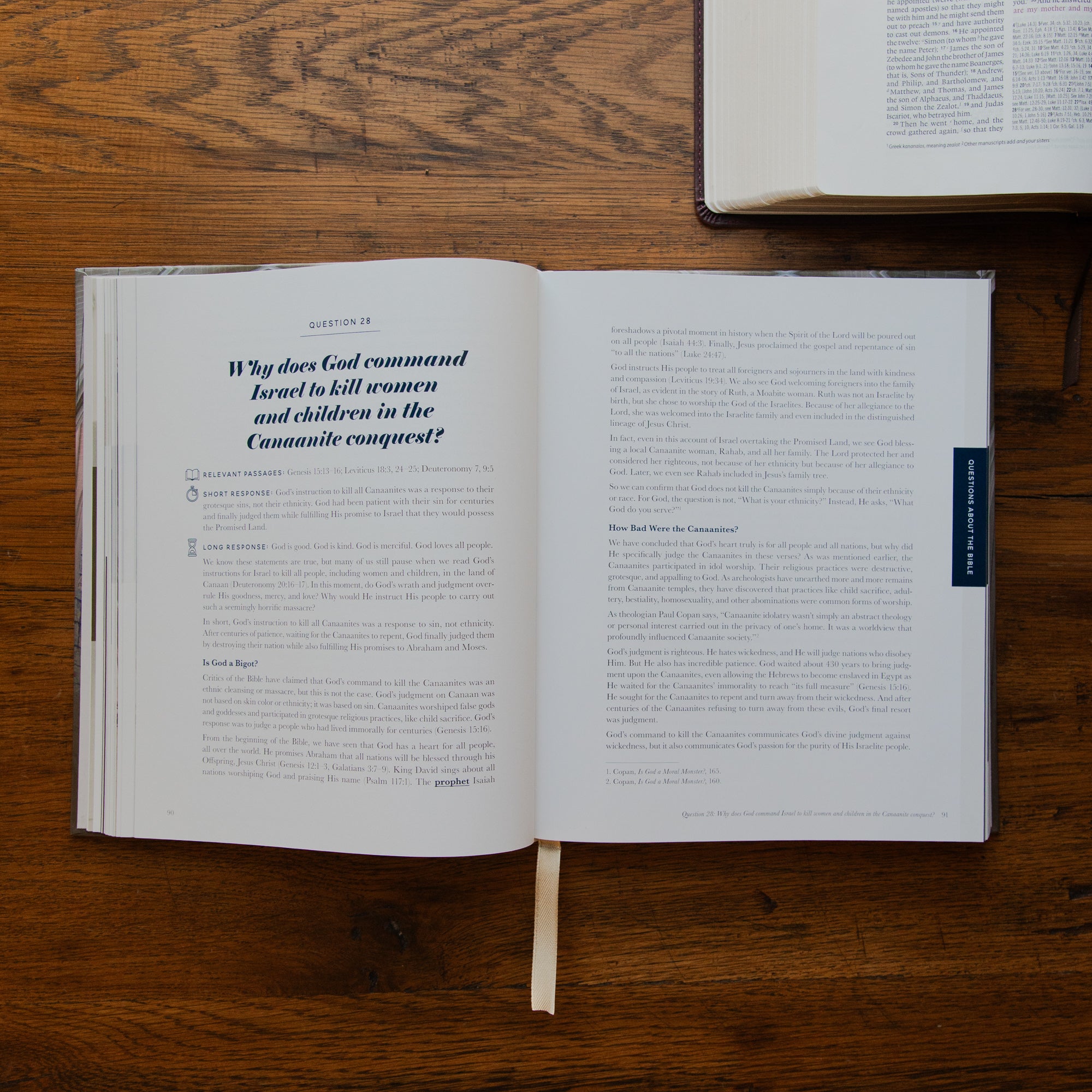 An open book lying on a wooden tabletop, showing printed text pages with a ribbon bookmark hanging from the center. Another book is partially visible at the top right.