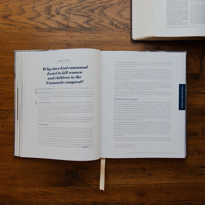 An open book lying on a wooden tabletop, showing printed text pages with a ribbon bookmark hanging from the center. Another book is partially visible at the top right.