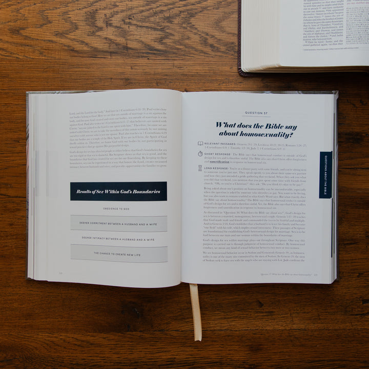 An open book laid flat on a wooden table, showing printed pages with text and a dark blue sidebar. Another book is partially visible at the top right.