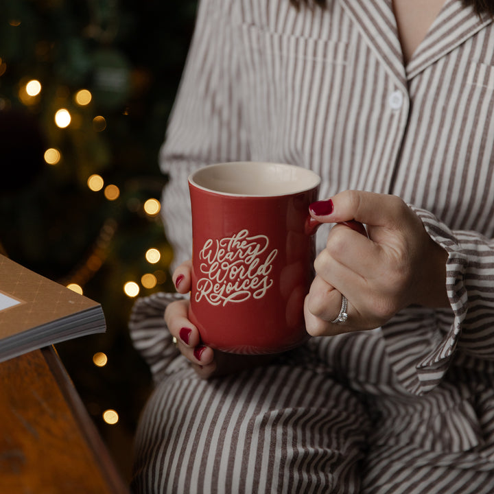 A person in brown and white striped pajamas holds a red ceramic mug with 'The World is Yours' written in white script, with a blurred Christmas tree and warm lights in the background.