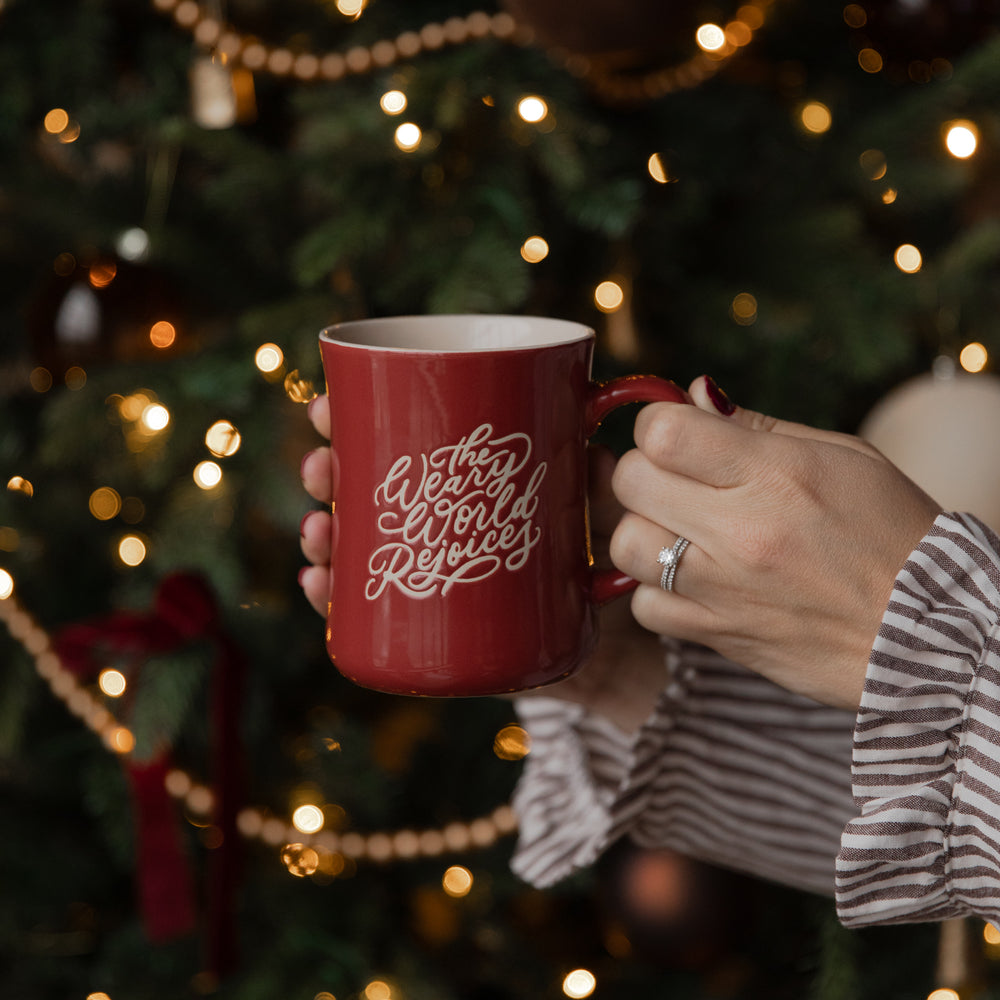 A person holds a red ceramic mug with 'The West Wing Reunion' written in white script in front of a blurred Christmas tree with warm lights.