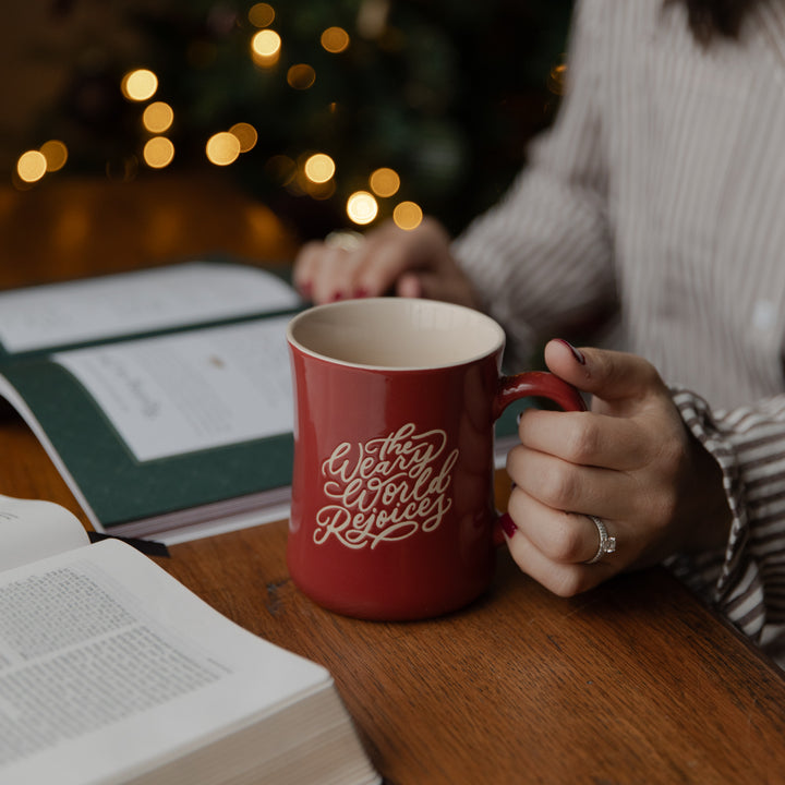 A person holds a red ceramic mug with white script text reading 'The World is Unending' on a wooden table, with an open book and warm string lights in the background.