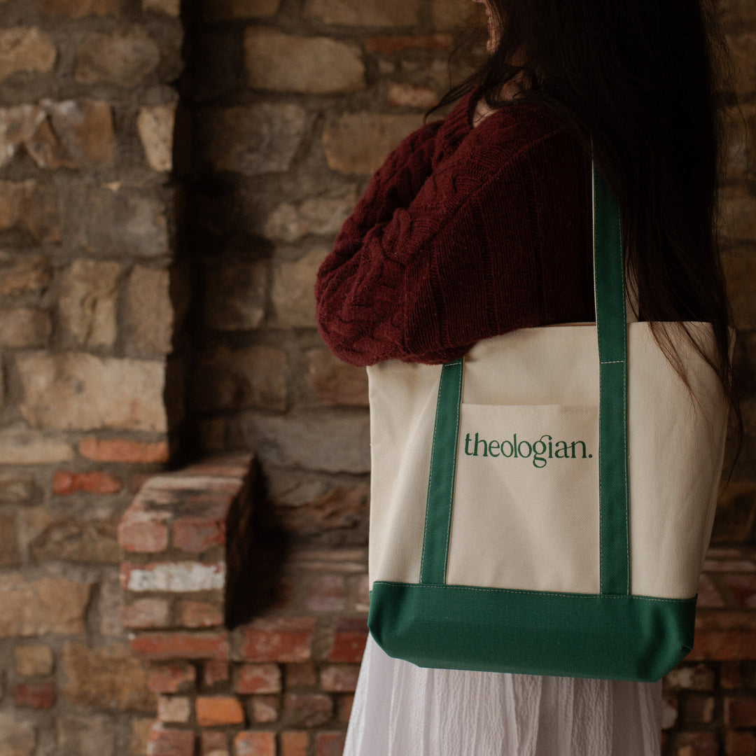 A woman with a red sweater and a white skirt holding a bag that says theologian on it over her shoulder near a brick wall