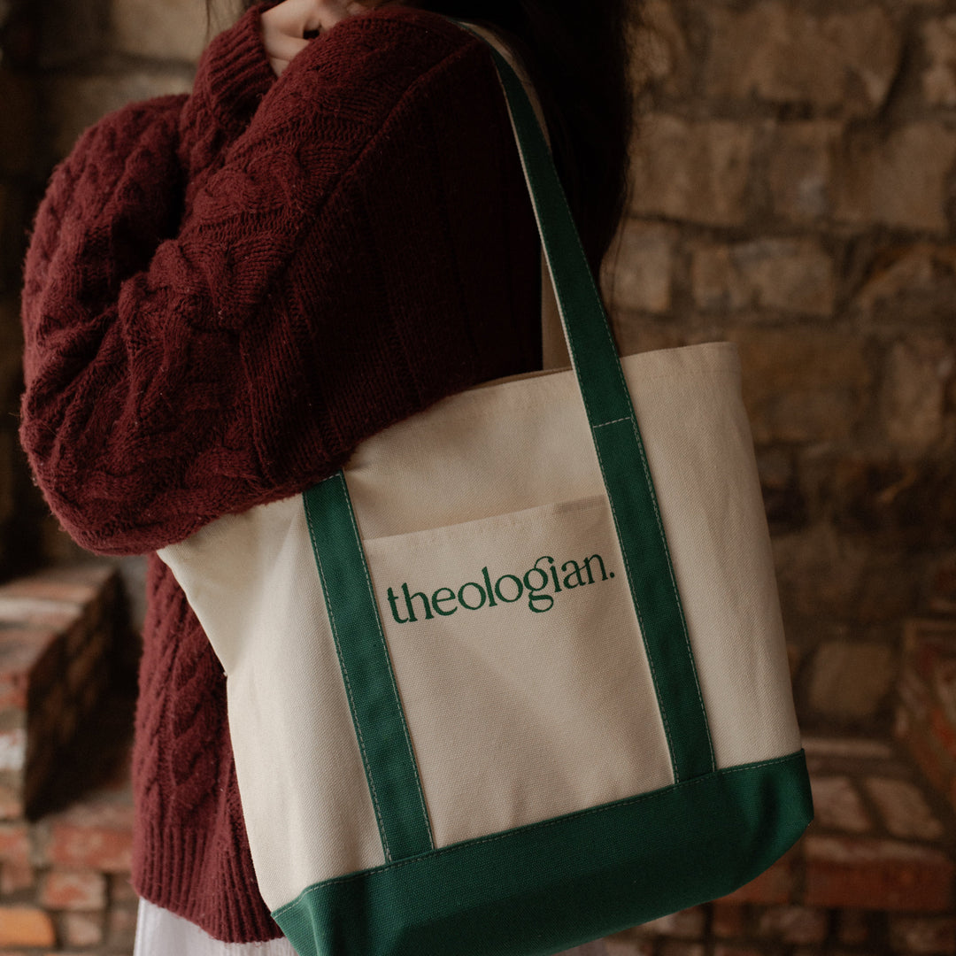 A woman with a red sweater holding a bag that says theologian on it over her shoulder with her arms crossed in front