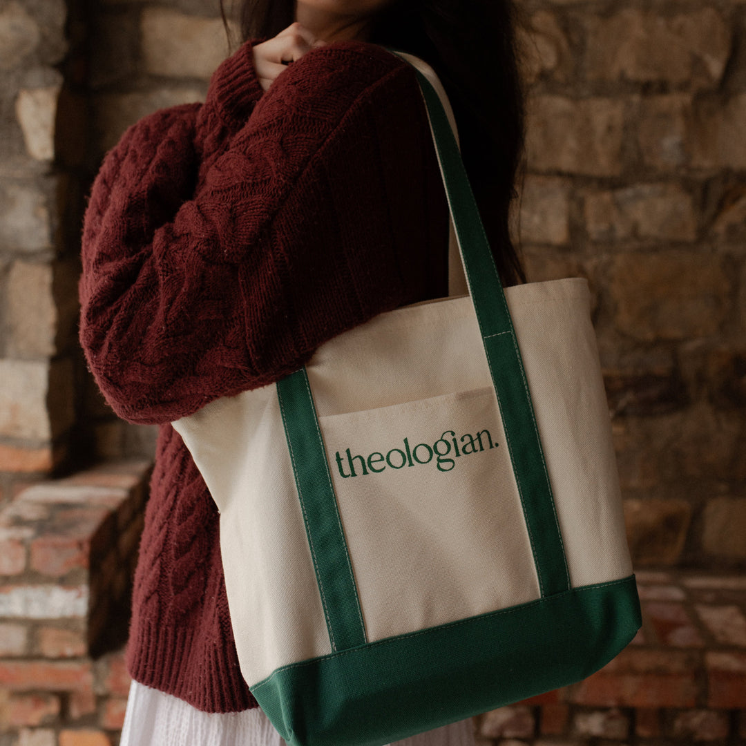 A woman with a red sweater holding a bag that says theologian on it over her shoulder while her arms are crossed in front of her