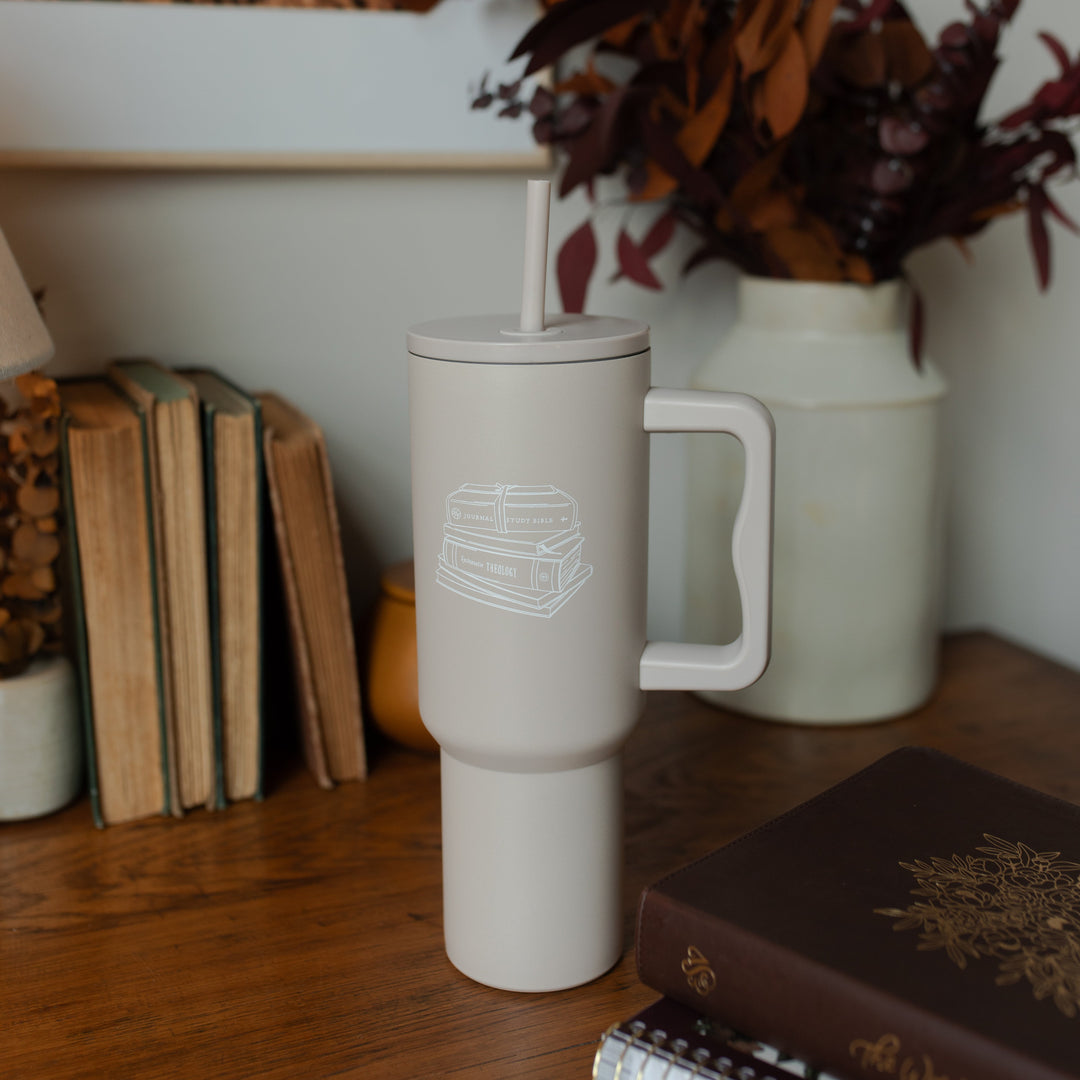 A taupe tumbler with an image of stacked books on it on a wooden desk near some plants