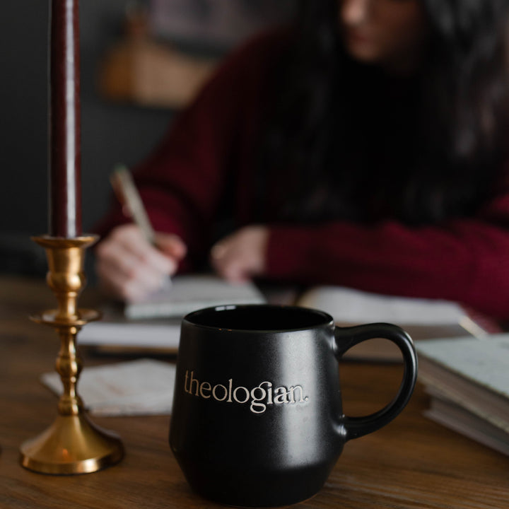 Black Theologian mug on table with woman writing in the background