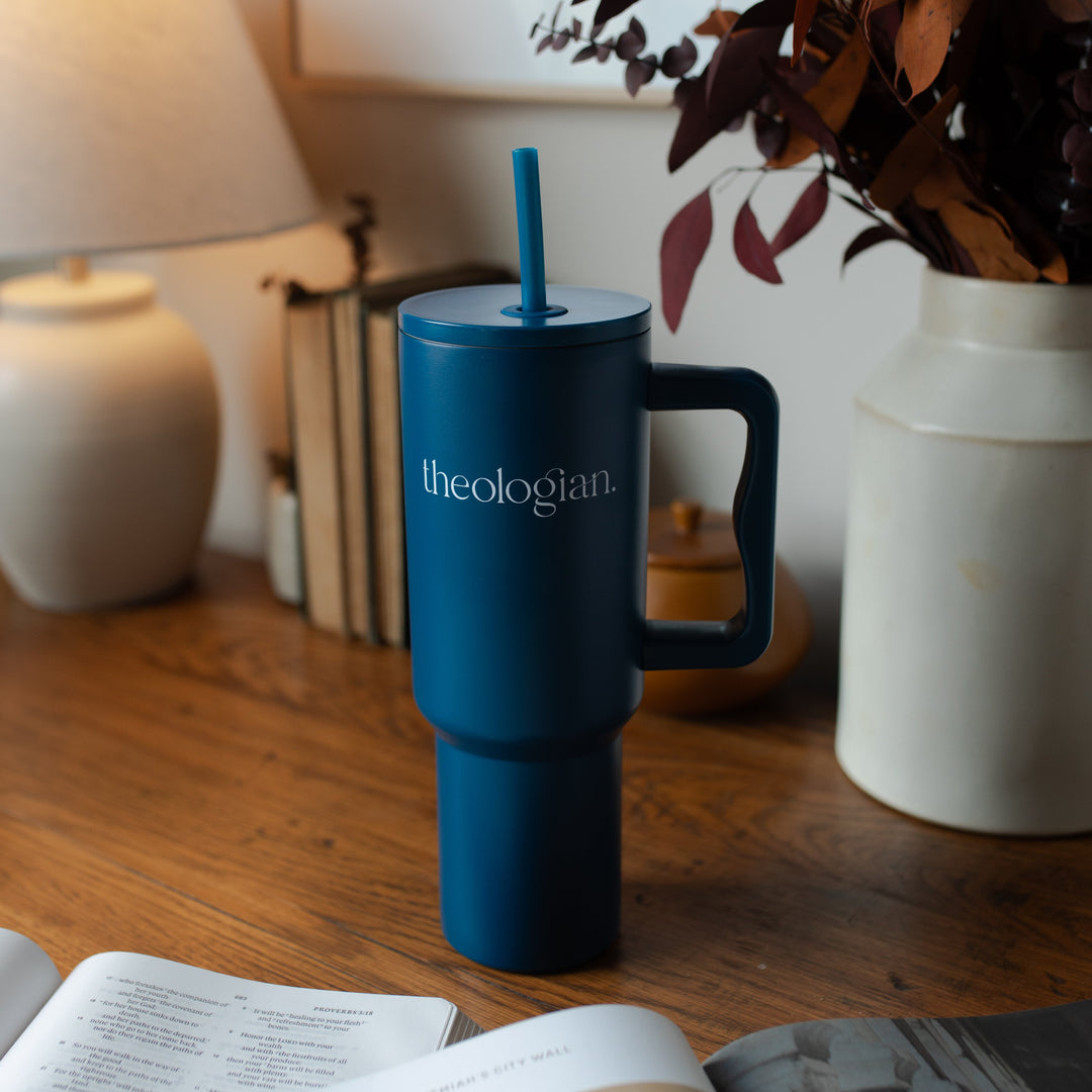 A blue tumbler with the words theologian on it on a wooden desk near a lamp and some plants