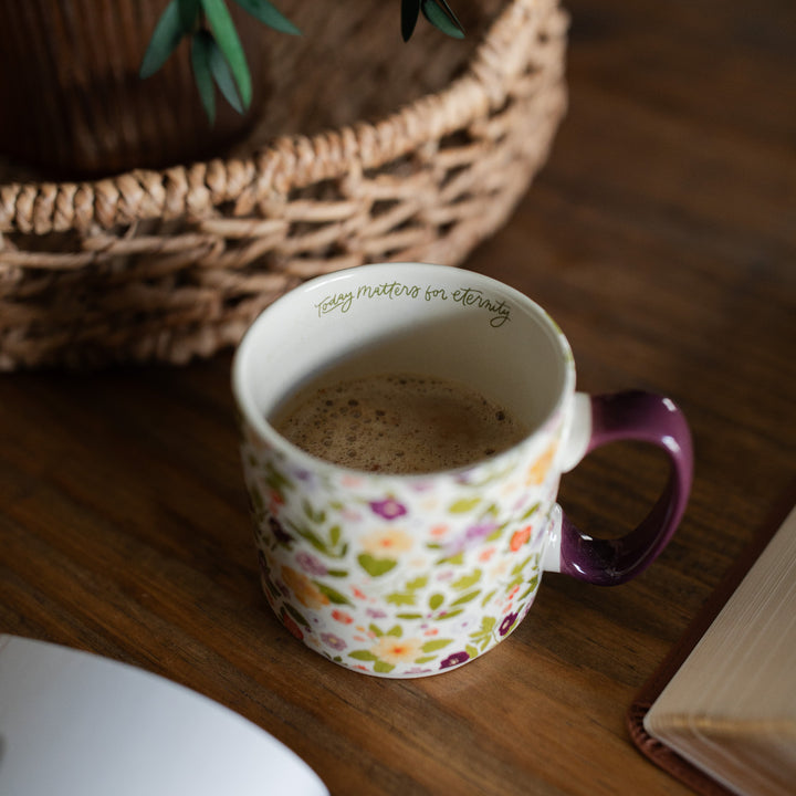 Over head shot of the Today Matters Magnolia Floral Mug on a brown wooden table next to a brown woven basket, and open Bible and an open book