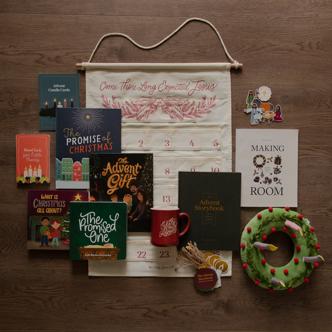 A Christmas advent arrangement laid flat on a dark wood surface, featuring a cream hanging banner with red text and numbers, several holiday books, a red mug, a green wreath with candles, small decorative items, and a white card reading 'Making a Room.'.