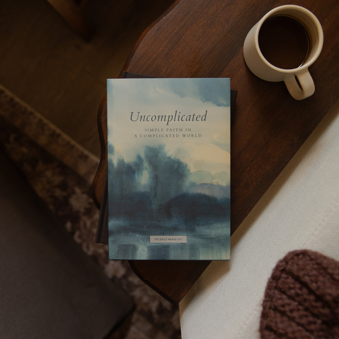 A book titled 'Uncomplicated: Simple Faith in a Complicated World' by Richard Swinburne lies on a dark wooden table next to a beige coffee mug. The book cover features a soft blue and gray abstract landscape.