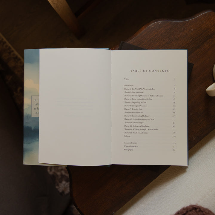 An open book laid on a dark wooden table, showing an interior table of contents page with a mostly white spread and a blue-toned scenic page at the left edge.