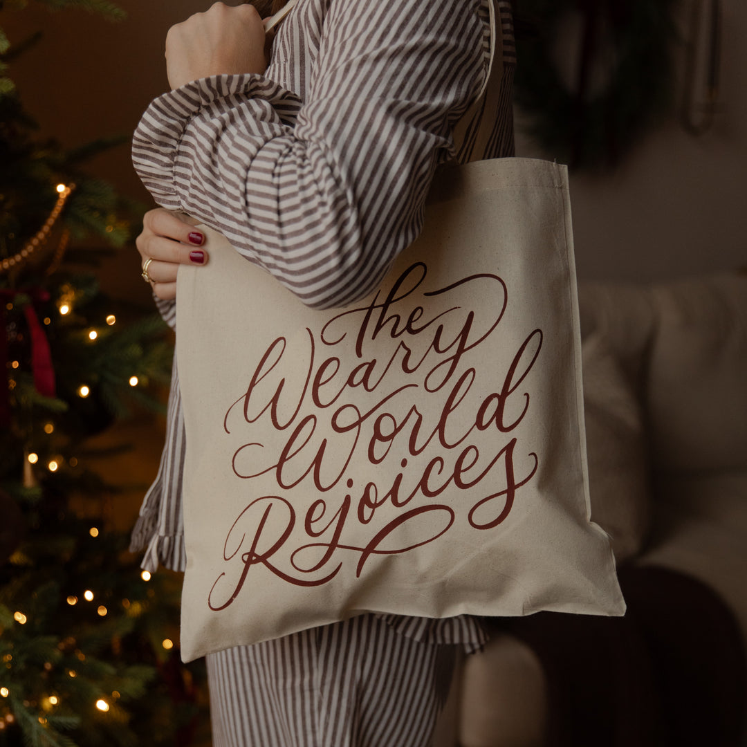 A person in a striped shirt holds a cream canvas tote bag with the phrase 'The Weary World Rejoices' in dark red script. The background is a cozy, dimly lit room with a blurred Christmas tree and couch.