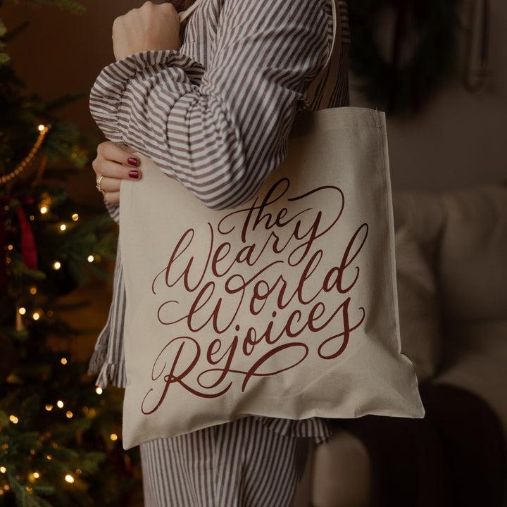 A person in a striped shirt holds a cream canvas tote bag with the phrase 'The Weary World Rejoices' in dark red script. The background is a cozy, dimly lit room with a blurred Christmas tree and couch.