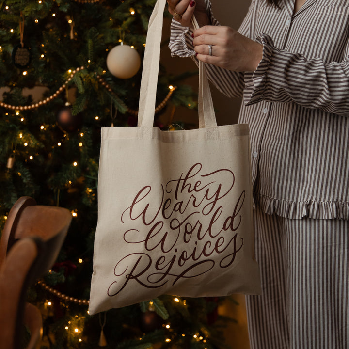 A person in a gray and white striped pajama set holds up a cream canvas tote bag with the phrase 'the Weary World Rejoices' printed in dark brown script, set against a cozy, warmly lit Christmas tree background.