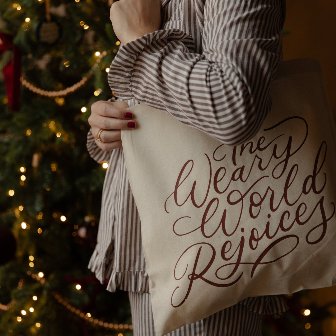 A person in a brown and white striped pajama set holds a cream canvas tote bag printed with the phrase 'Merry World Rejoices' in dark brown cursive script, set against a warm, blurred Christmas tree background.