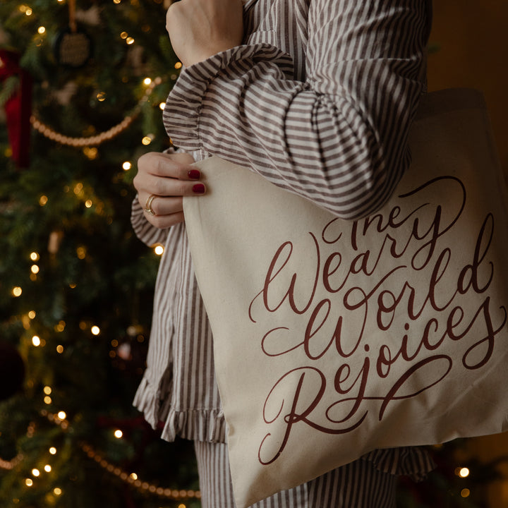 A person in a brown and white striped pajama set holds a cream canvas tote bag printed with the phrase 'Merry World Rejoices' in dark brown cursive script, set against a warm, blurred Christmas tree background.