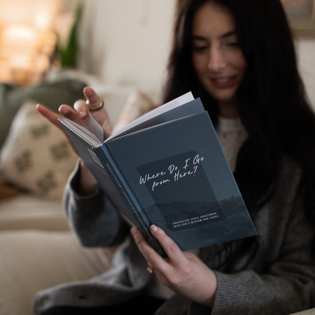 A woman sitting on a white couch, reading from a book named Where Do I Go from Here, flipping through pages