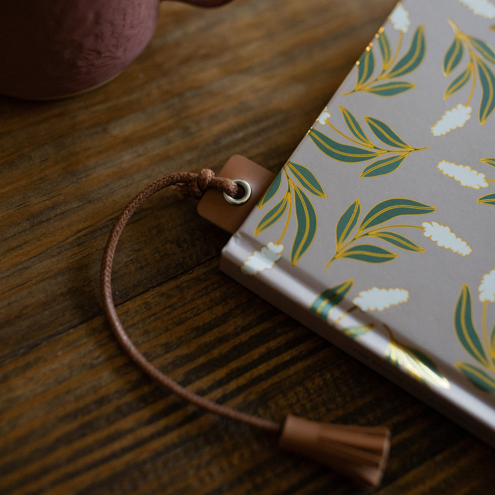 One Brown You Belong Here Leatherette Bookmark inside a book laying on a wooden table next to a coffee cup