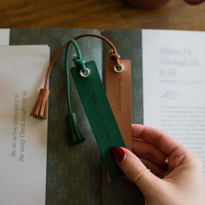 A woman's hand holding You Belong Here Leatherette Bookmarks over a bible study that is laying on a wooden table