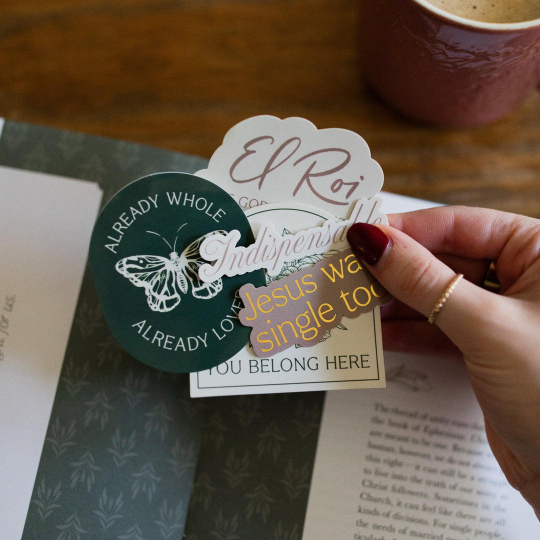 A woman holding various stickers in the You Belong Here Sticker Pack over an open bible study that is laying on a wooden table next to a coffee cup