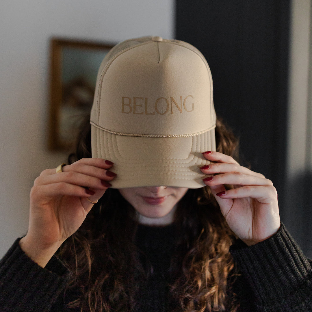 A Woman holding a You belong Here Trucker Hat by the tip while it sits on her head