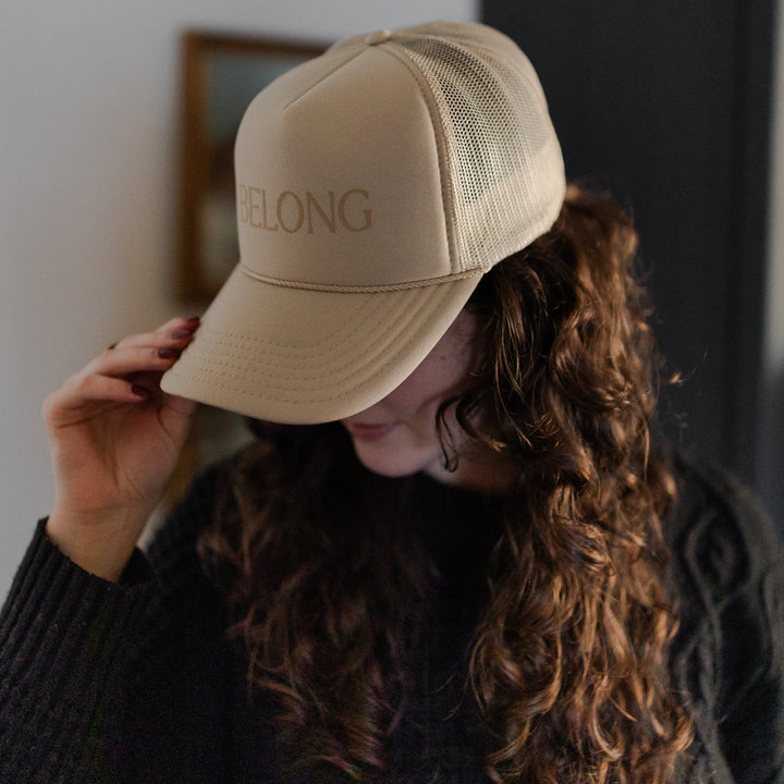 A woman wearing a You Belong Here Trucker Hat looking to the side with one hand holding the tip