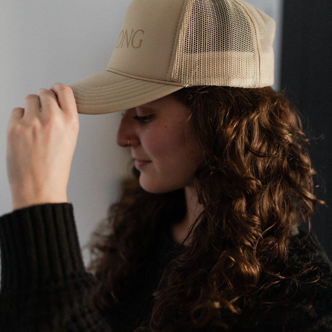 A Woman wearing a You Belong Here Trucker Hat looking to the side and down while she has one hand on the tip of the hat