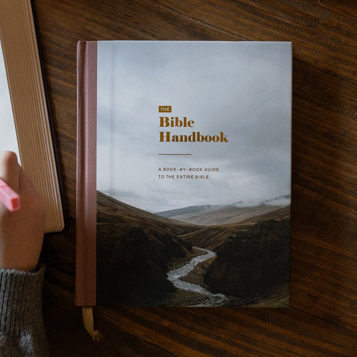 A Bible Handbook book lies open on a wooden table, with a hand holding a pink highlighter beside it. A woven basket and part of another open book are visible in the background.